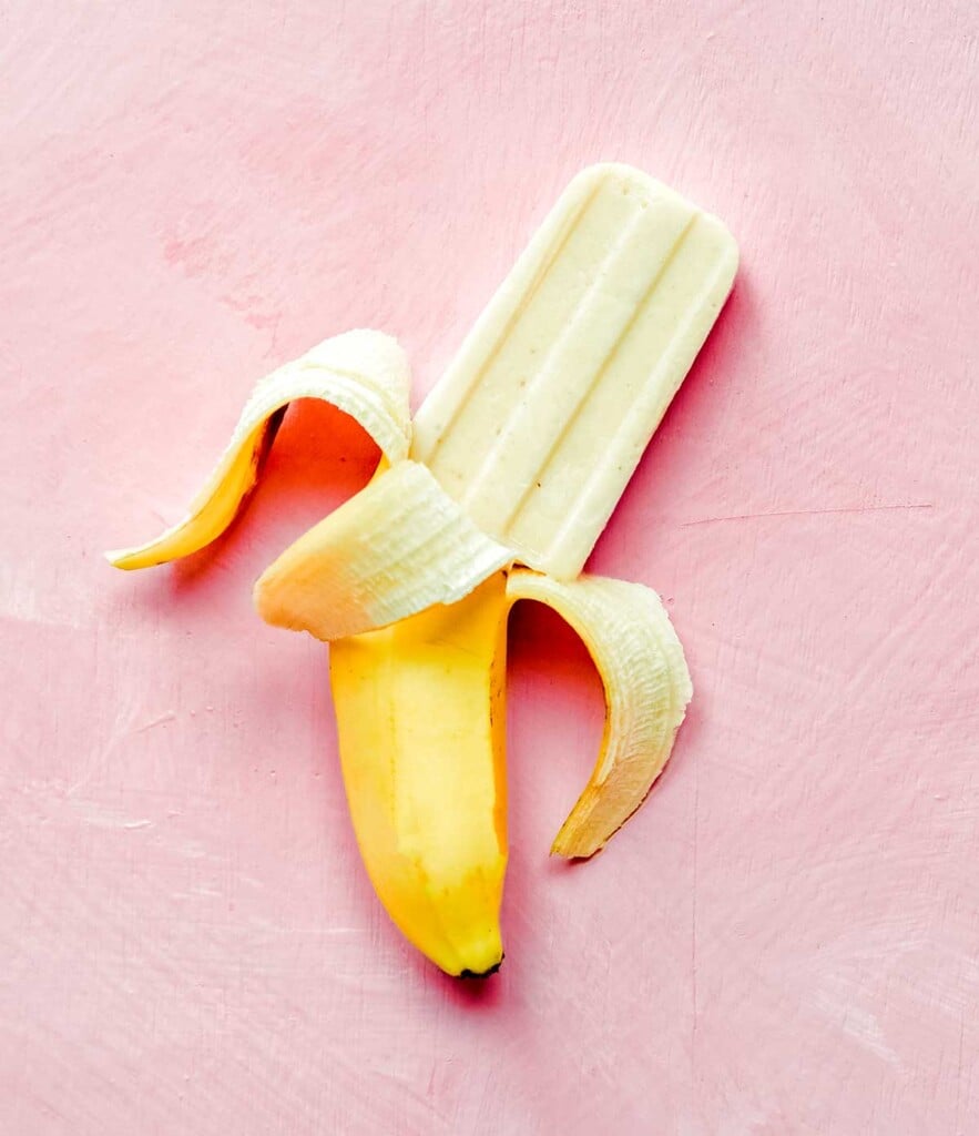 A banana popsicles and a banana peel laid out on a pink background