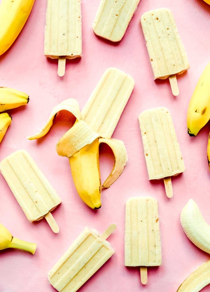 A bunch of banana popsicles and banana peels laid out on a pink background