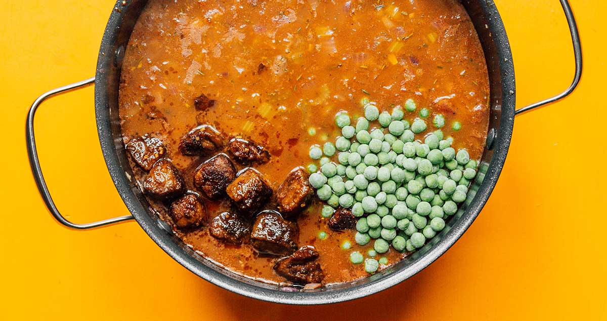 A large pot filled with vegetarian beef stew ingredients, including cooked beefless tips and frozen peas