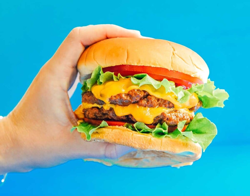 A hand holding out a double seitan burger on a fluffy bun with tomato, lettuce, and cheese