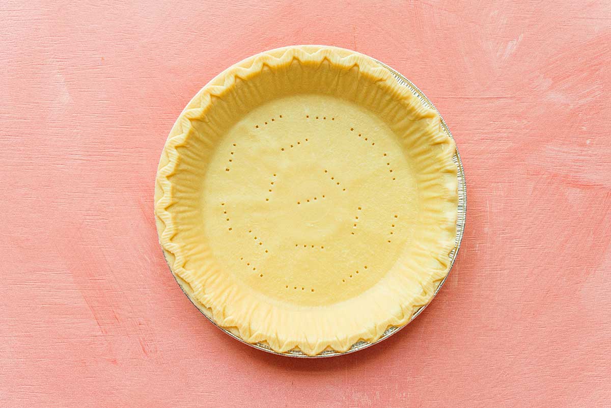 An uncooked, empty pie crust in a tin cooking tray on a light pink background