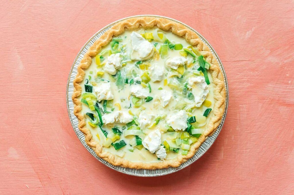 A cooked pie crust filled with sliced leeks and goat cheese pieces with an egg mixture poured on top