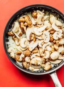 A frying pan holds the beginnings of a delicious mushroom stroganoff, with sliced mushrooms and onions set against a vibrant red background.