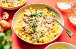 A bowl of pasta topped with creamy mushroom stroganoff sauce, garnished with parsley. A fork rests on the edge, surrounded by mushrooms and glasses on a red surface.