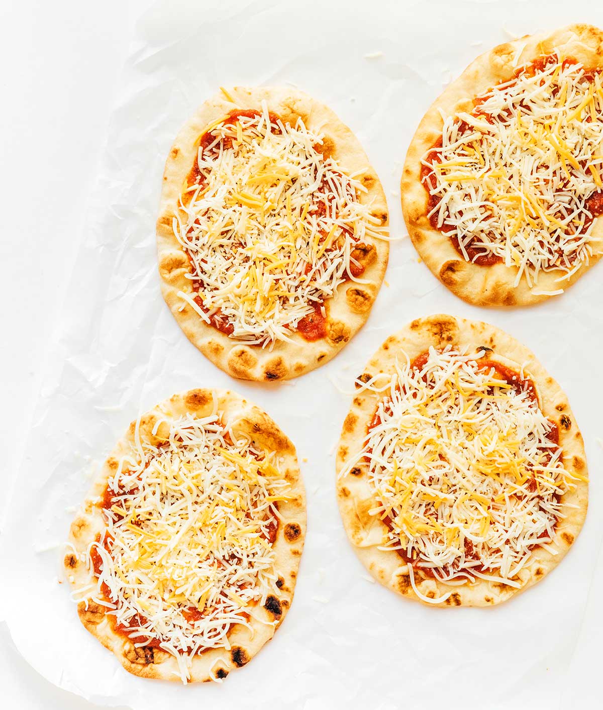 Four uncooked naan pizzas topped with sauce, mozzarella, and cheddar cheese