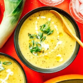 A bowl of potato leek soup garnished with sliced green onions and cream, accompanied by a wooden spoon, rests on a red surface with fresh leek and chilled ice water nearby.