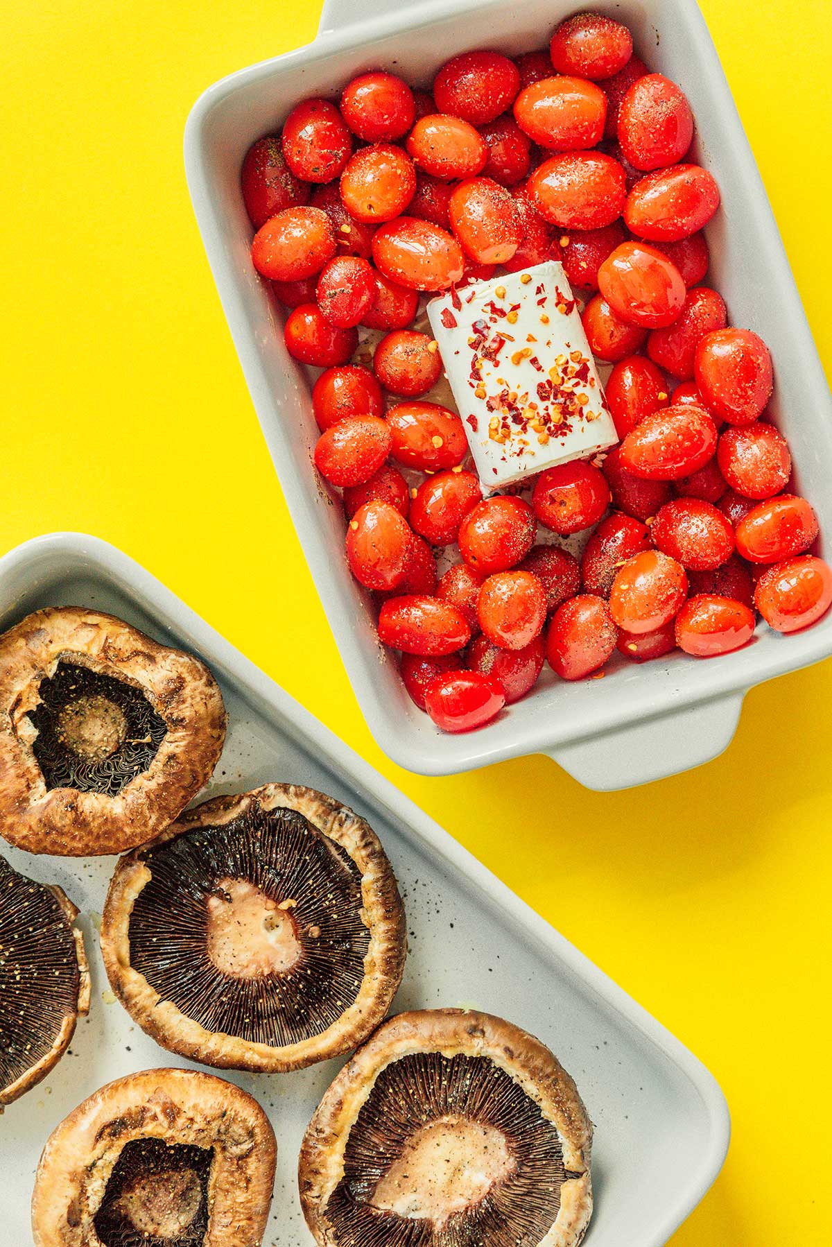 A casserole dish filled with uncooked cherry tomatoes and goat cheese beside a baking tray filled with portobello mushrooms