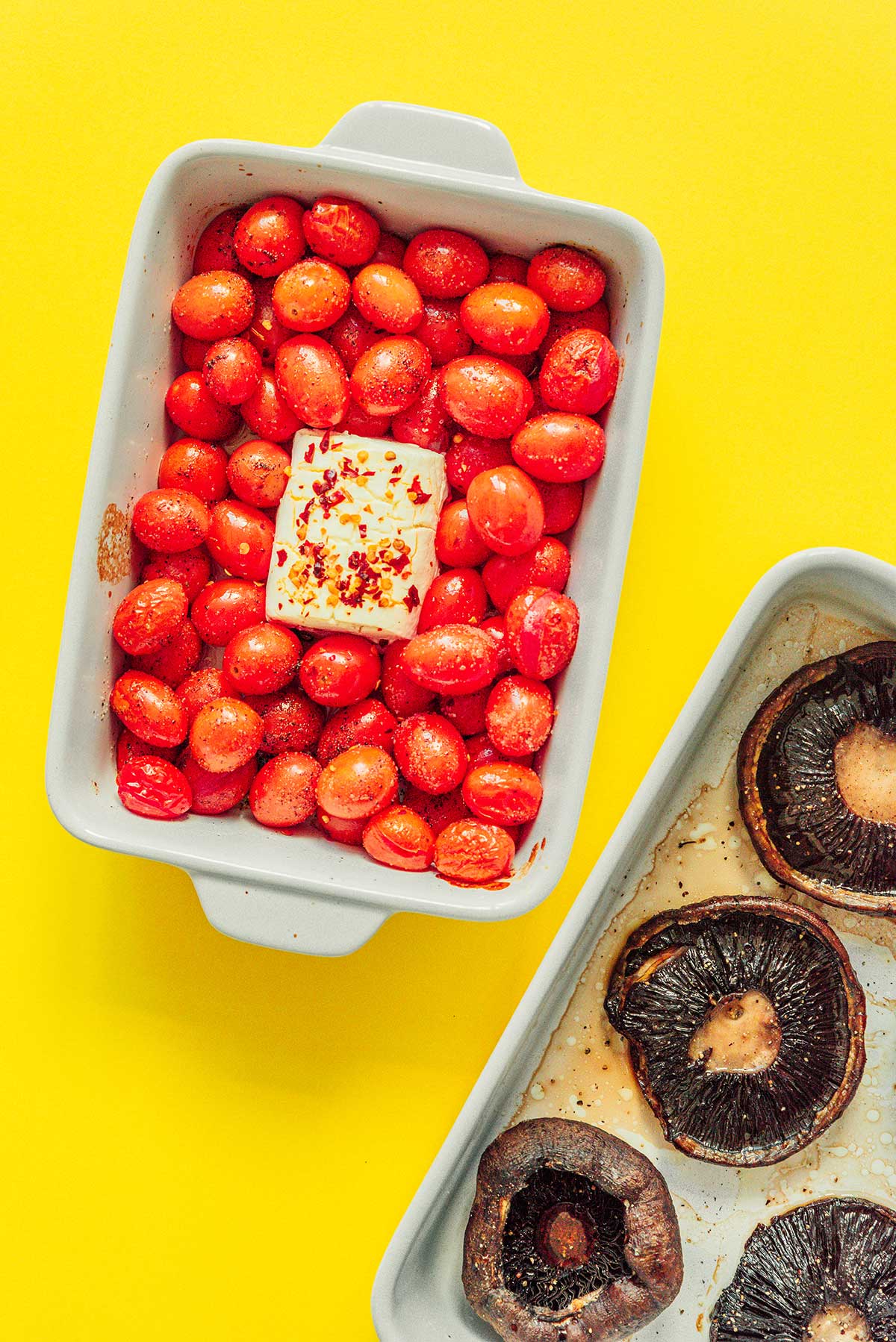 A casserole dish filled with baked cherry tomatoes and goat cheese beside a baking tray filled with baked portobello mushrooms