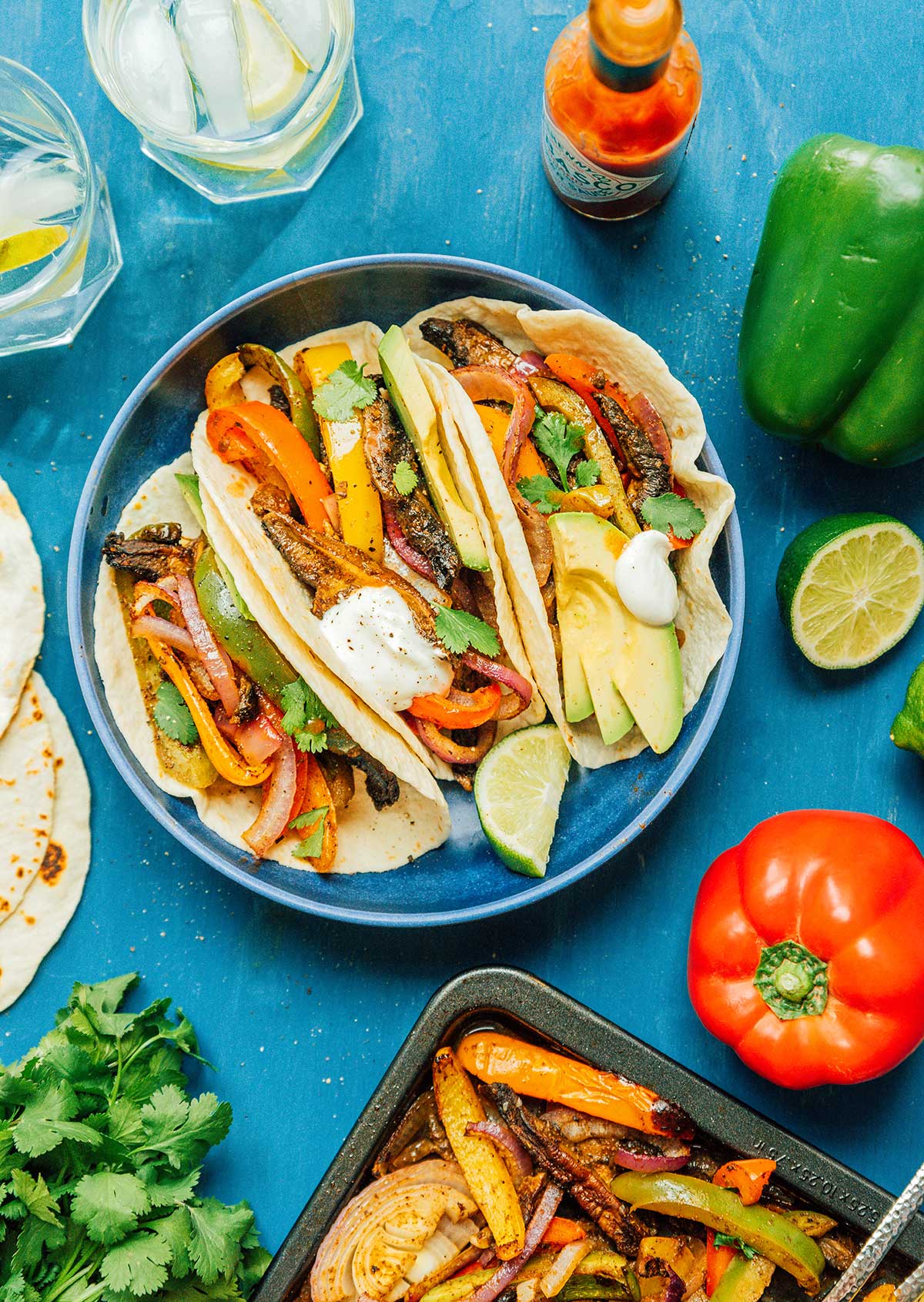 A blue plate filled with 3 vegetable fajitas and surrounded by various fajita ingredients
