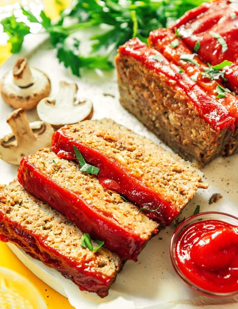Three slices of mushroom meatloaf on a white plate surrounded by a dipping bowl of ketchup and other ingredients