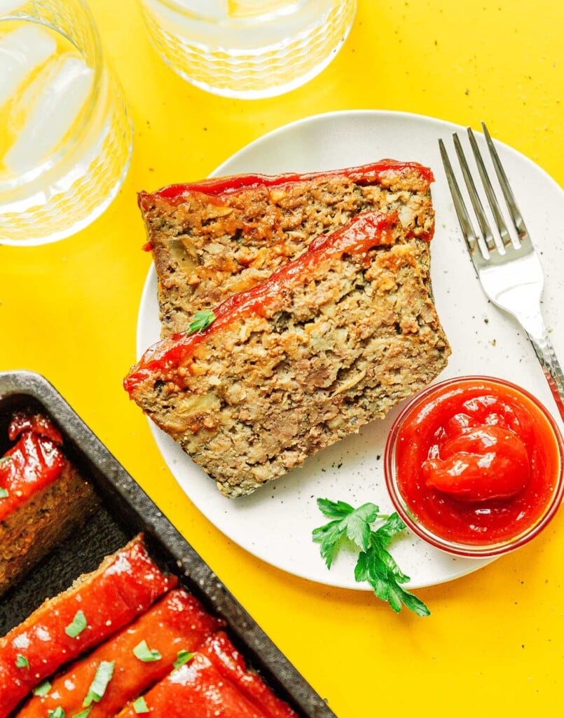 Two slices of mushroom meatloaf on a white plate along with a metal fork and a dipping bowl of ketchup