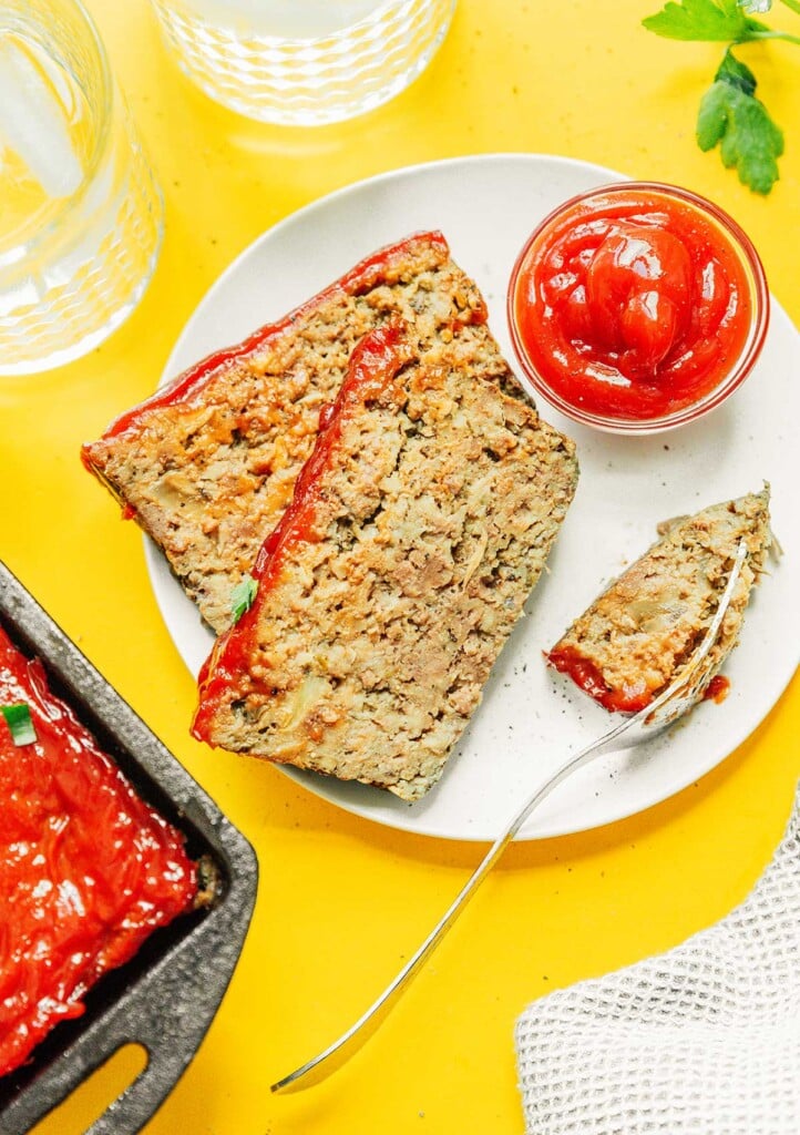 Two slices of mushroom meatloaf on a white plate along with a metal fork and dipping bowl of ketchup