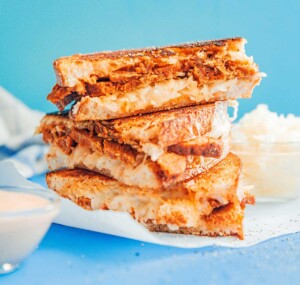 Stacked grilled sandwiches reminiscent of a vegetarian Reuben are filled with pulled pork and sauerkraut on white paper. A small bowl of sauce sits beside them against a blue background.