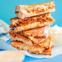 Stack of grilled cheese sandwiches with a creamy, vegetarian Reuben-style filling, set on white parchment paper against a blue background.