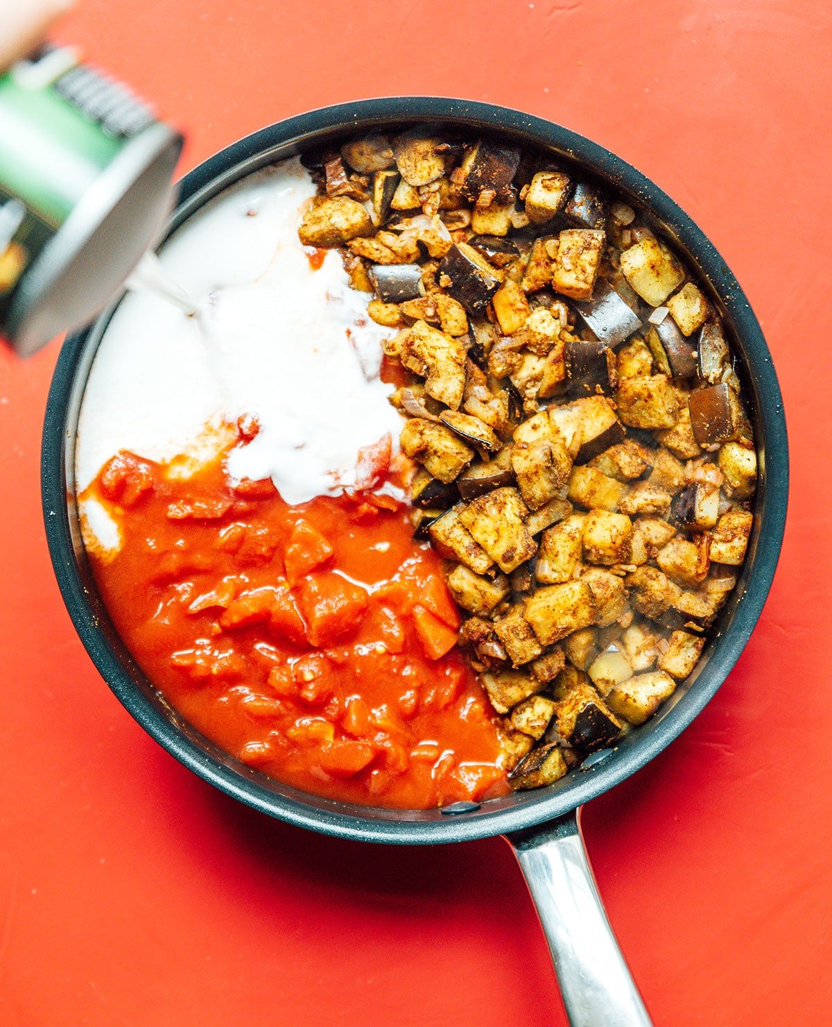 A skillet filled with diced eggplant, shallots, and garlic, along with a can of coconut milk and a can of diced tomatoes