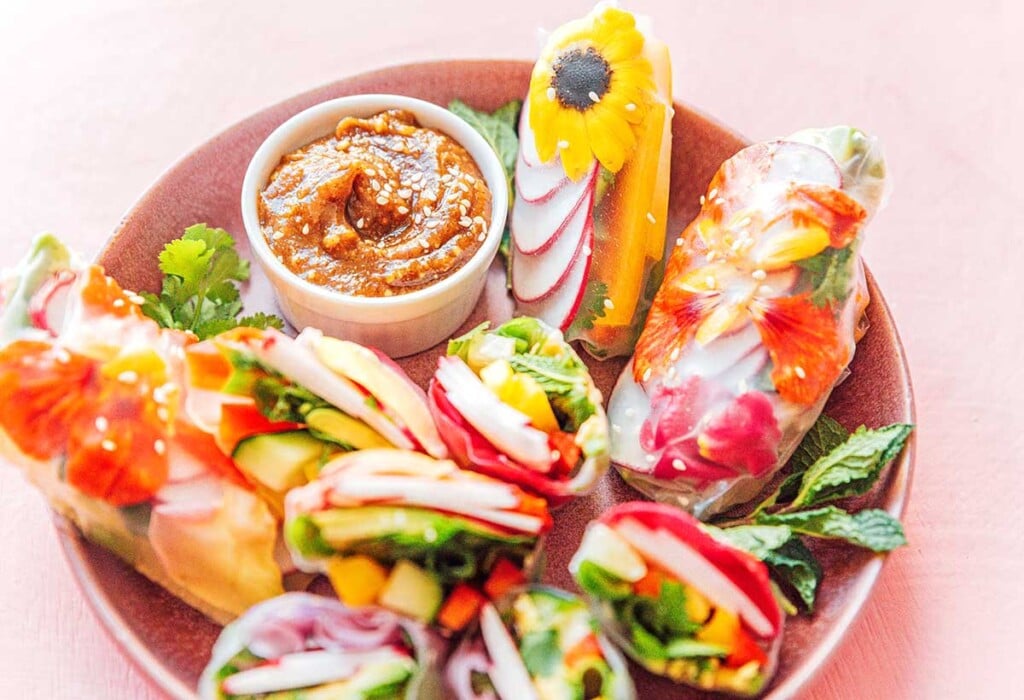 A pink bowl filled with nine edible flower spring rolls and a smaller bowl of almond butter dipping sauce
