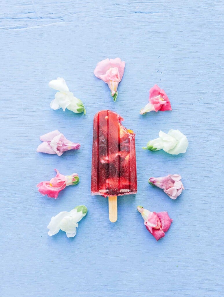 A hibiscus popsicle with a bite taken out laying on a blue background and surrounded by pink, purple, and white hibiscus flowers
