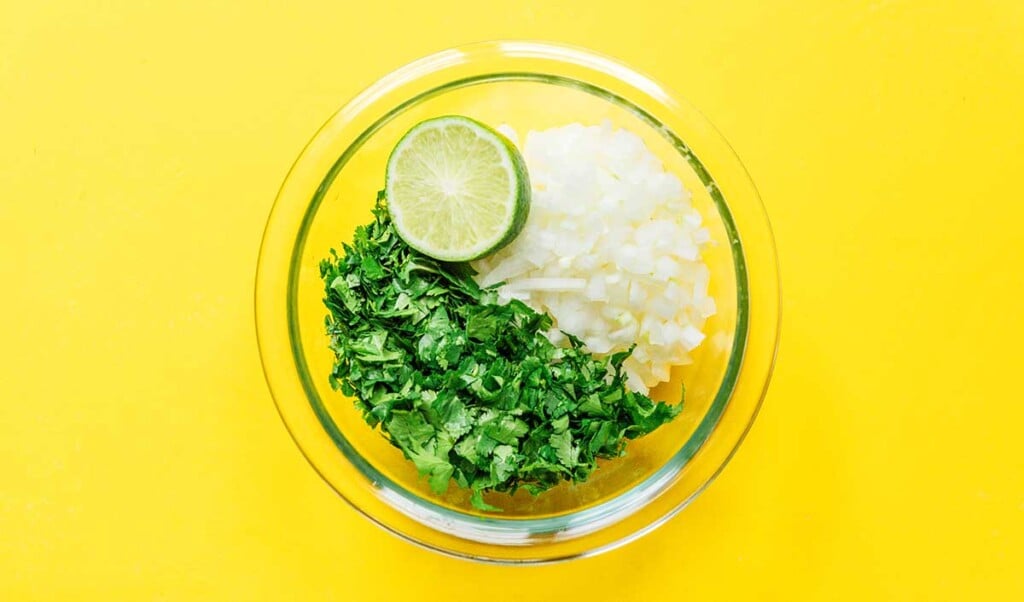 A clear glass bowl filled with chopped cilantro, chopped onion, and half of a lime.