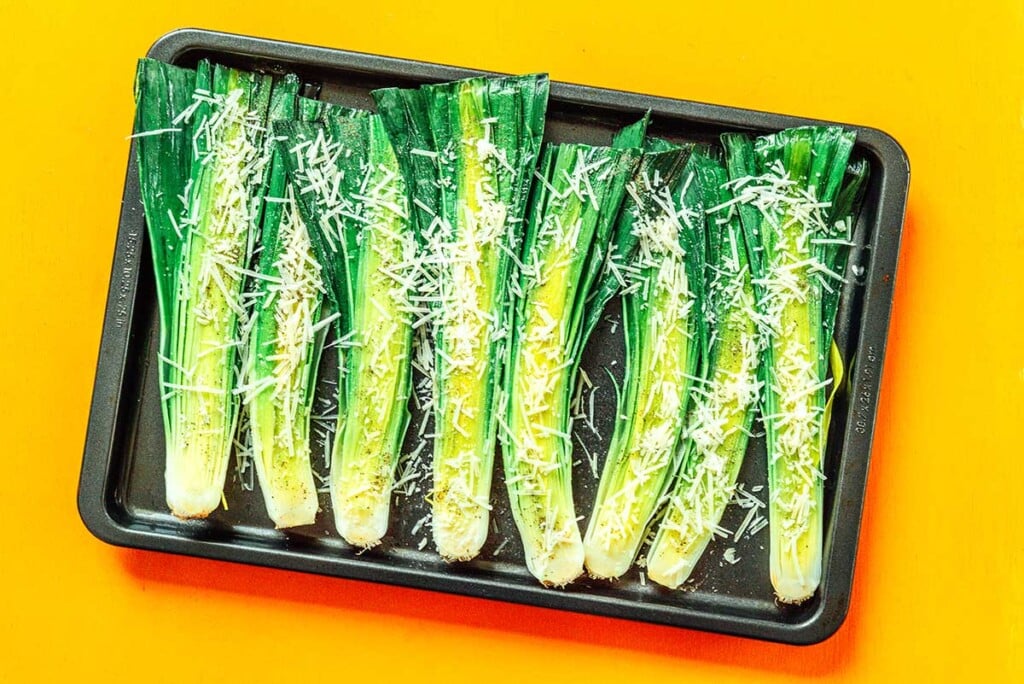 A baking sheet filled with a row of eight uncooked, seasoned leek halves laying cutside up