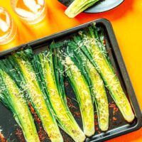 Roasted leeks topped with grated cheese rest invitingly on a baking tray, accompanied by drinks and an extra serving of these delightful roasted leeks on a plate.