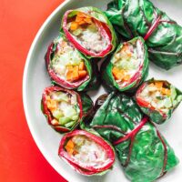 A white plate showcases chard wraps, each filled with sliced carrots, cucumbers, and rice, against a vibrant red backdrop.