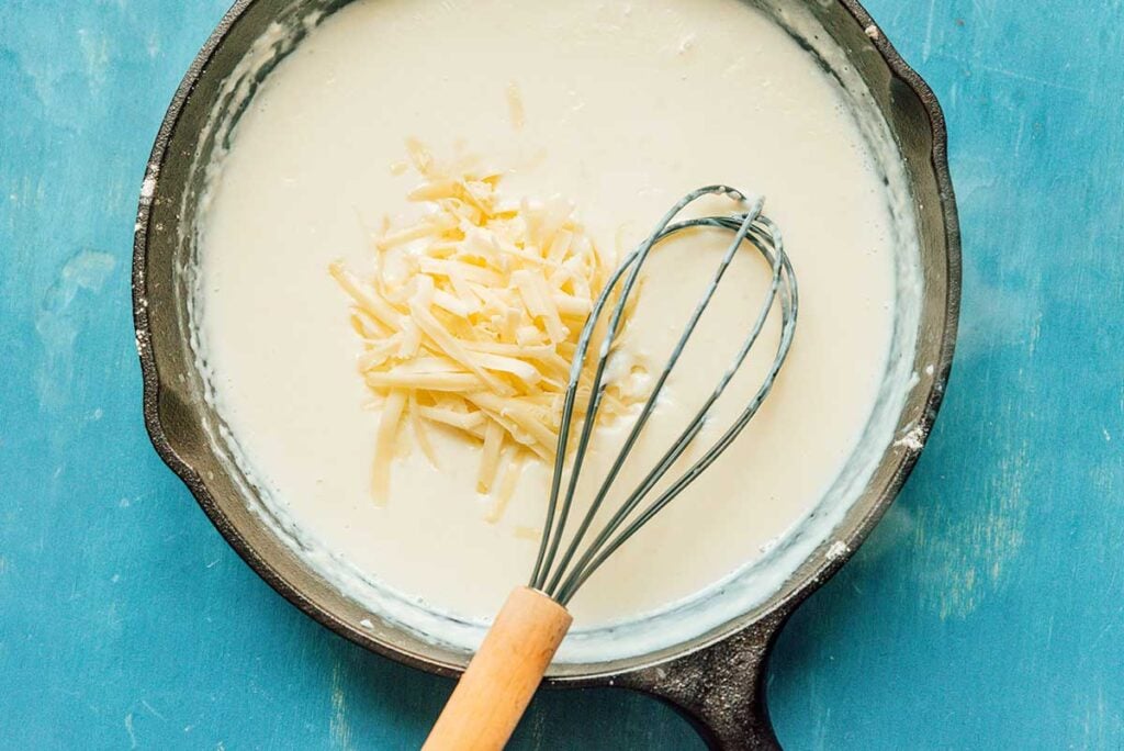 A whisk stirring the bechamel sauce ingredients in a cast iron skillet