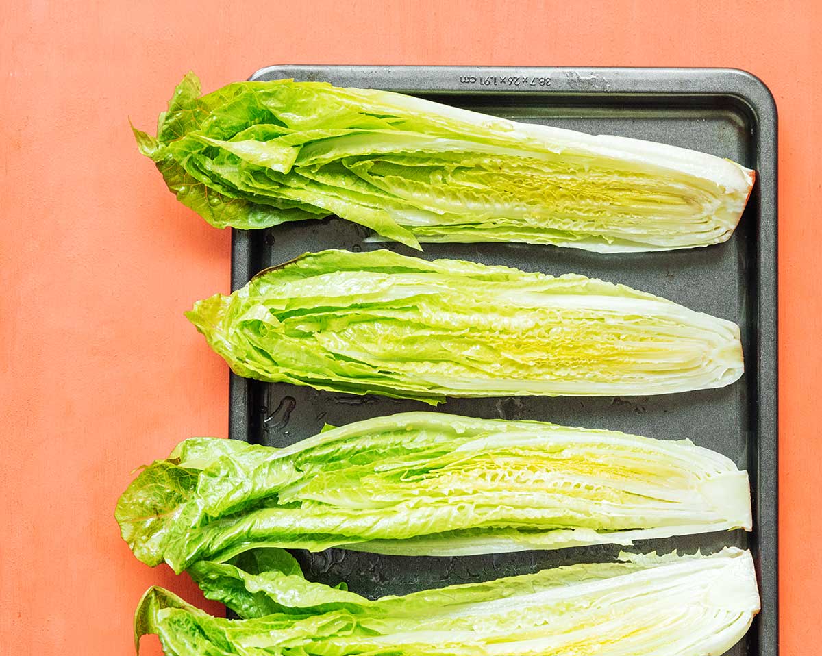 Four uncooked romaine heart halves lined up cut side-up on a baking tray
