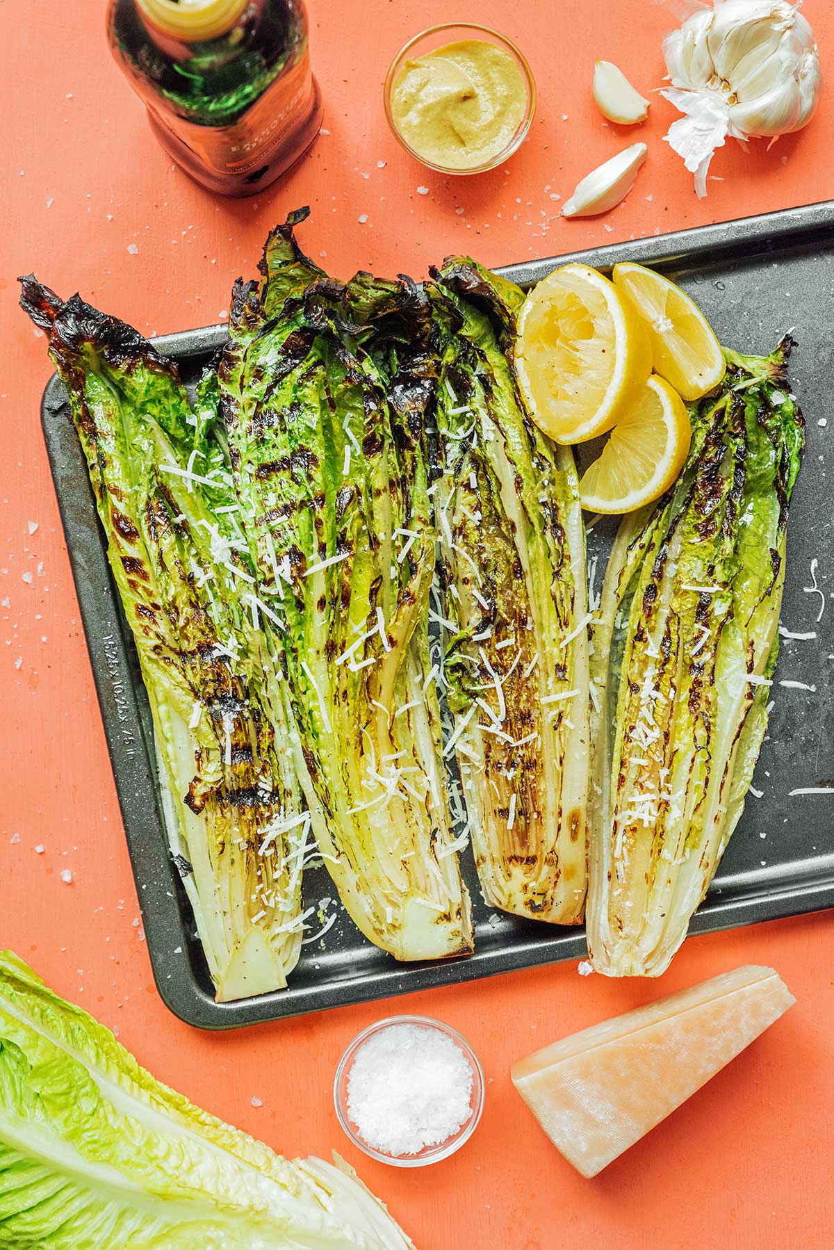 Four grilled romaine heart halves topped with parmesan cheese sitting atop a baking sheet next to lemon slices