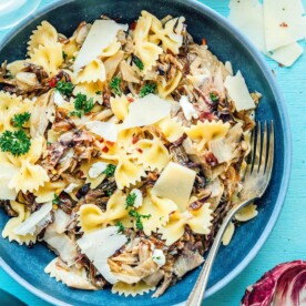 A bowl of radicchio pasta adorned with thin cheese slices and a parsley garnish. A fork rests on the plate, while cheese chunks are scattered in the background on a vibrant blue surface.