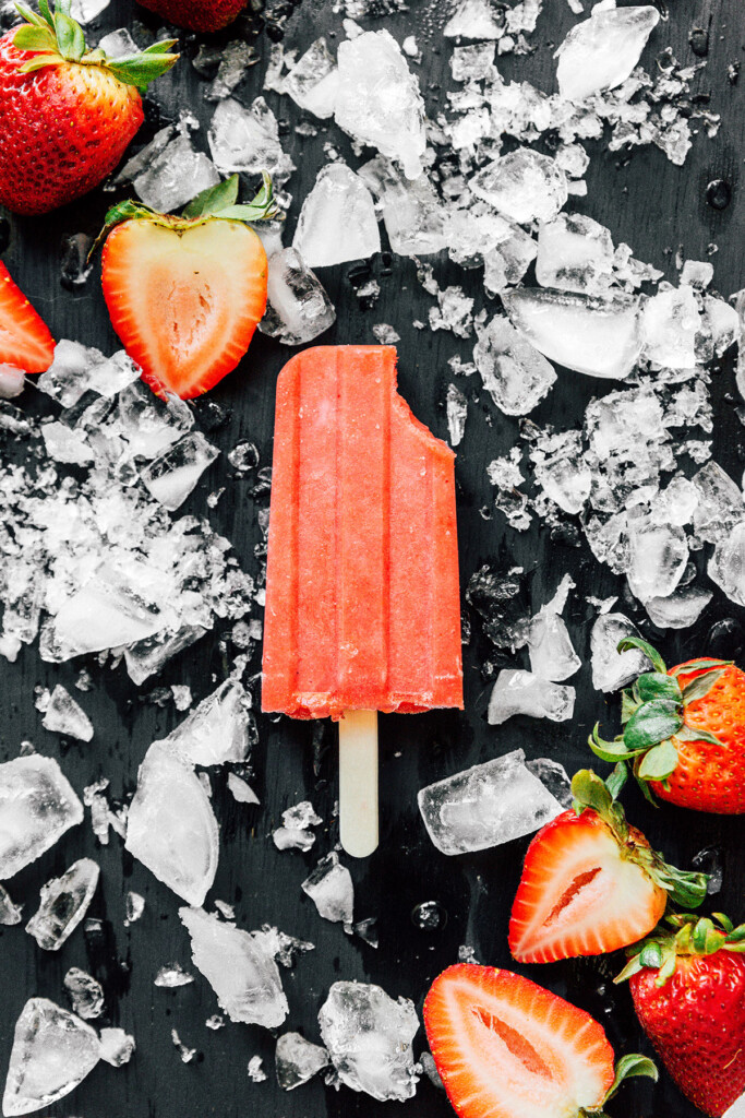 A sugar-free strawberry popsicle with a bite taken out on a black background and surrounded by ice and strawberries