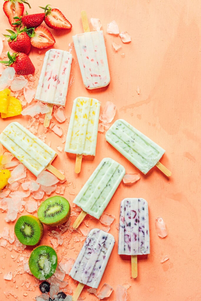 Various 3 ingredient fruit and yogurt popsicles arranged on an orange background and surrounded by ice, strawberries, pineapple, kiwis, and blueberries