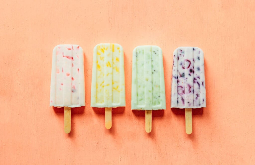 Strawberry, pineapple, kiwi, and blueberry popsciles lined up on an orange background