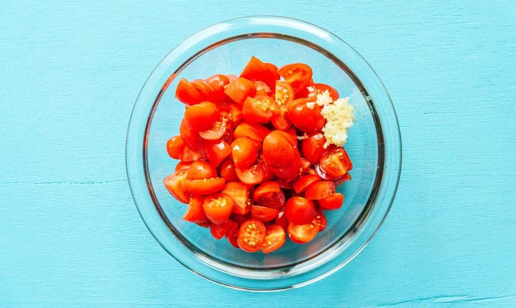 A clear glass bowl filled with quartered cherry tomatoes and minced garlic