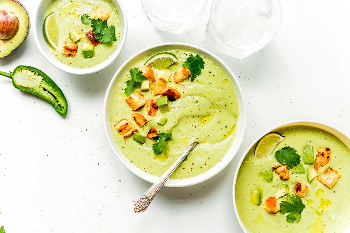 Three white bowls filled with vegan avocado soup and topped with a lime wedge, croutons, and cilantro