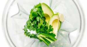 Ingredients in a blender: cilantro, avocado slices, garlic clove, green bell pepper pieces, and a pinch of salt—perfect for crafting a smooth and creamy avocado soup.