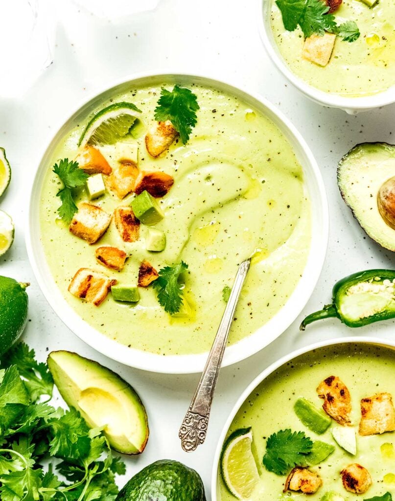 A white bowl filled with creamy avocado soup and topped with a lime wedge, diced avocado, croutons, and cilantro