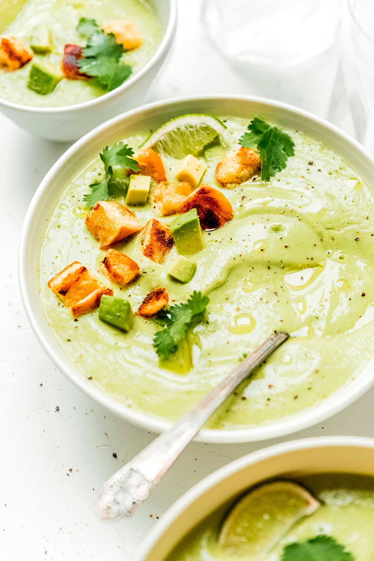 A white bowl filled with vegan avocado soup and topped with a lime wedge, diced avocado, croutons, and cilantro