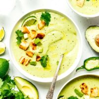 Bowls of creamy avocado soup garnished with croutons, avocado cubes, and cilantro, surrounded by lime wedges, jalapeño slices, avocado halves, and fresh cilantro leaves on a white background.