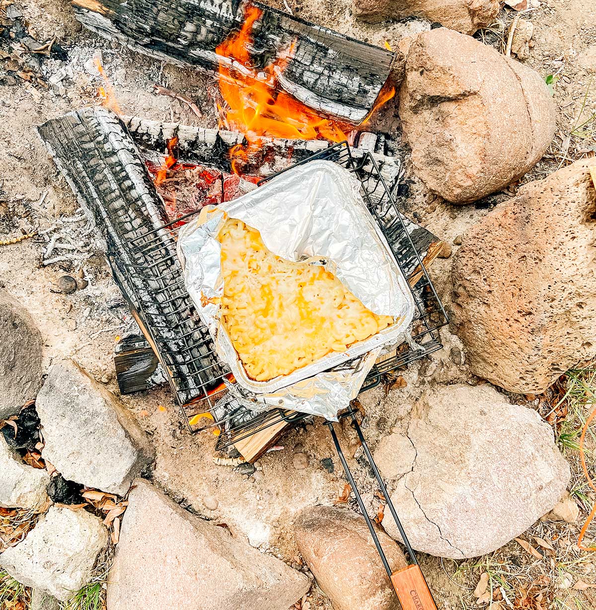 Cooking a disposable aluminum container of campfire mac n cheese on logs over a campfire