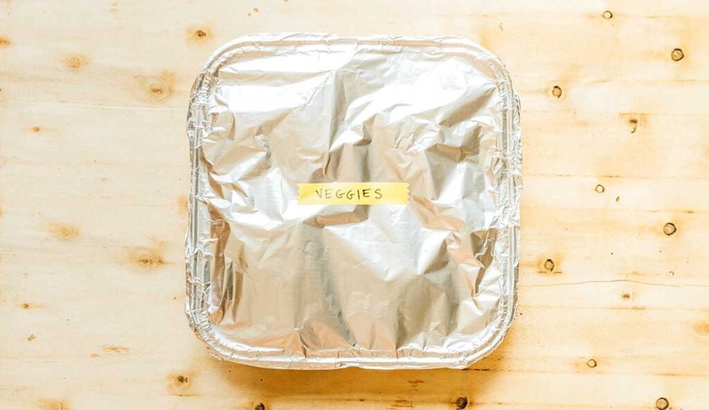 An aluminum container filled with uncooked roasted veggies, covered with foil, and labeled "veggies"