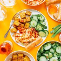 A vibrant spread of vegan Korean BBQ delights graces the yellow table, featuring plates of tofu, cucumber salad, kimchi, and spring rolls, complemented by glasses of iced water and an array of condiments.