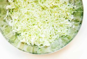 Shredded cabbage sits in a metal mixing bowl on a white surface, ready to embark on its transformation journey. Discover how to make sauerkraut with ease as you turn these humble ingredients into a tangy delight.