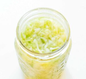 An open jar filled with chopped cabbage, likely sauerkraut, sits invitingly on a white background. Curious about its tangy allure? Discover how to make sauerkraut and bring this classic delight into your kitchen.
