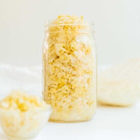 A jar of finely chopped homemade sauerkraut is filled to the brim, with a small bowl of sauerkraut beside it on a light background.