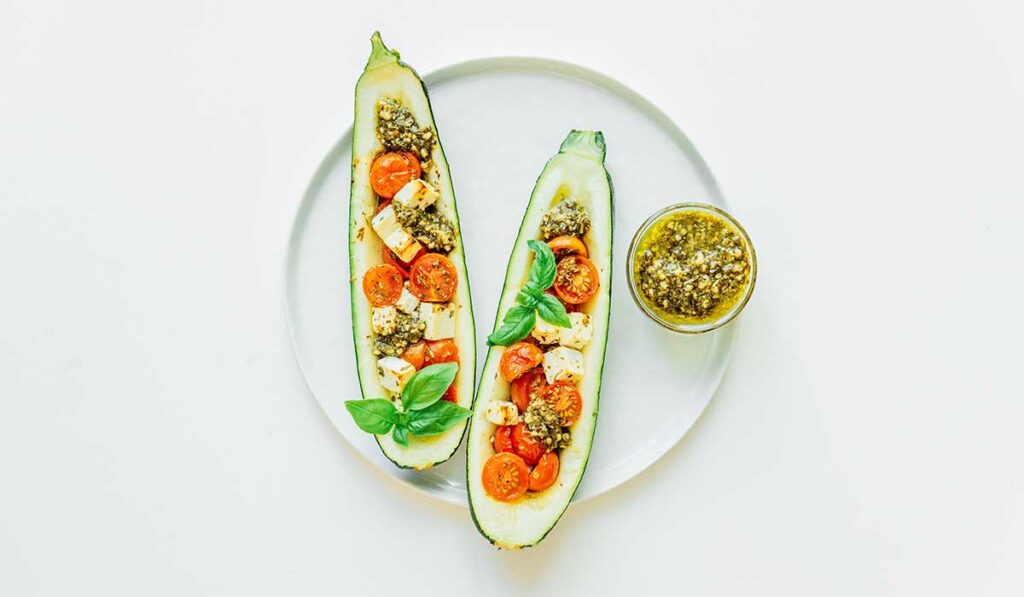 Two Mediterranean stuffed zucchini boats on a white plate with a small bowl of fresh pesto