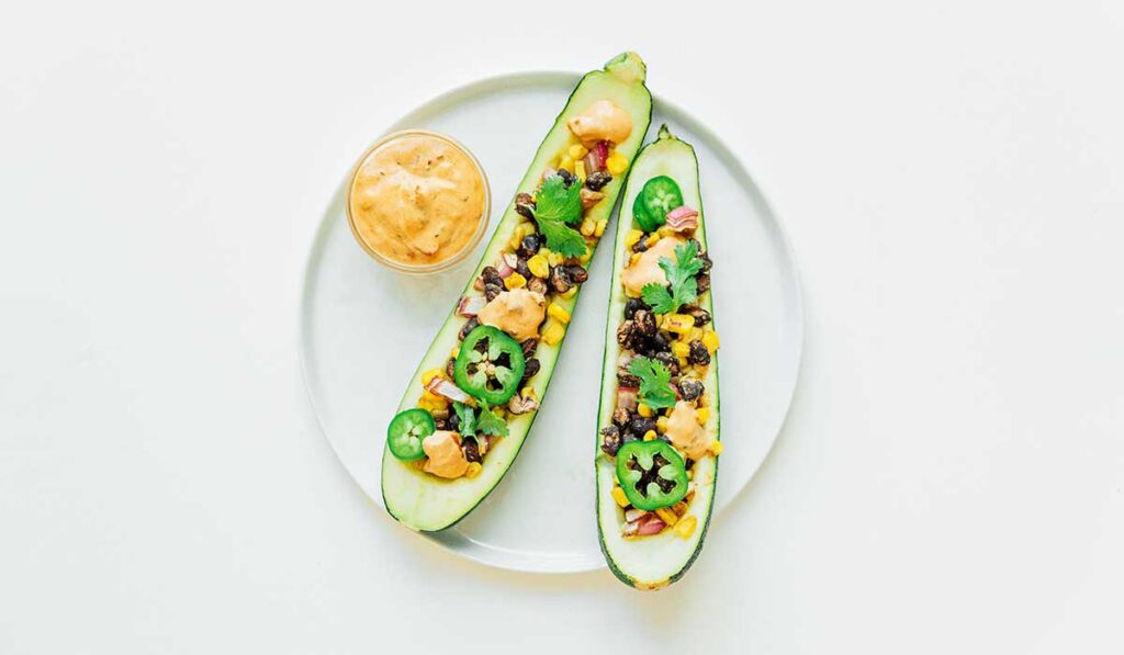 Two Mexican stuffed zucchini boats on a white plate with a small bowl of creamy adobo sauce