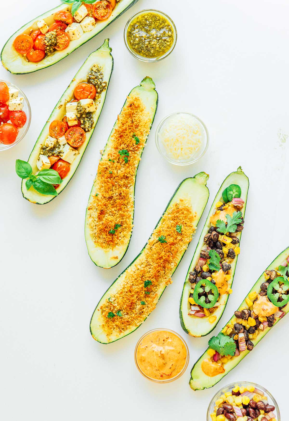 Six stuffed zucchini halves arranged on a white background and surrounded by small bowls of various toppings