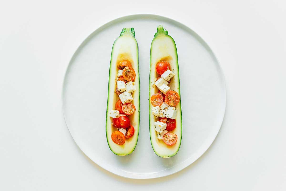 Two Mediterranean zucchini boats on a white plate filled with cherry tomatoes, feta cheese, Italian seasoning, salt, and pepper