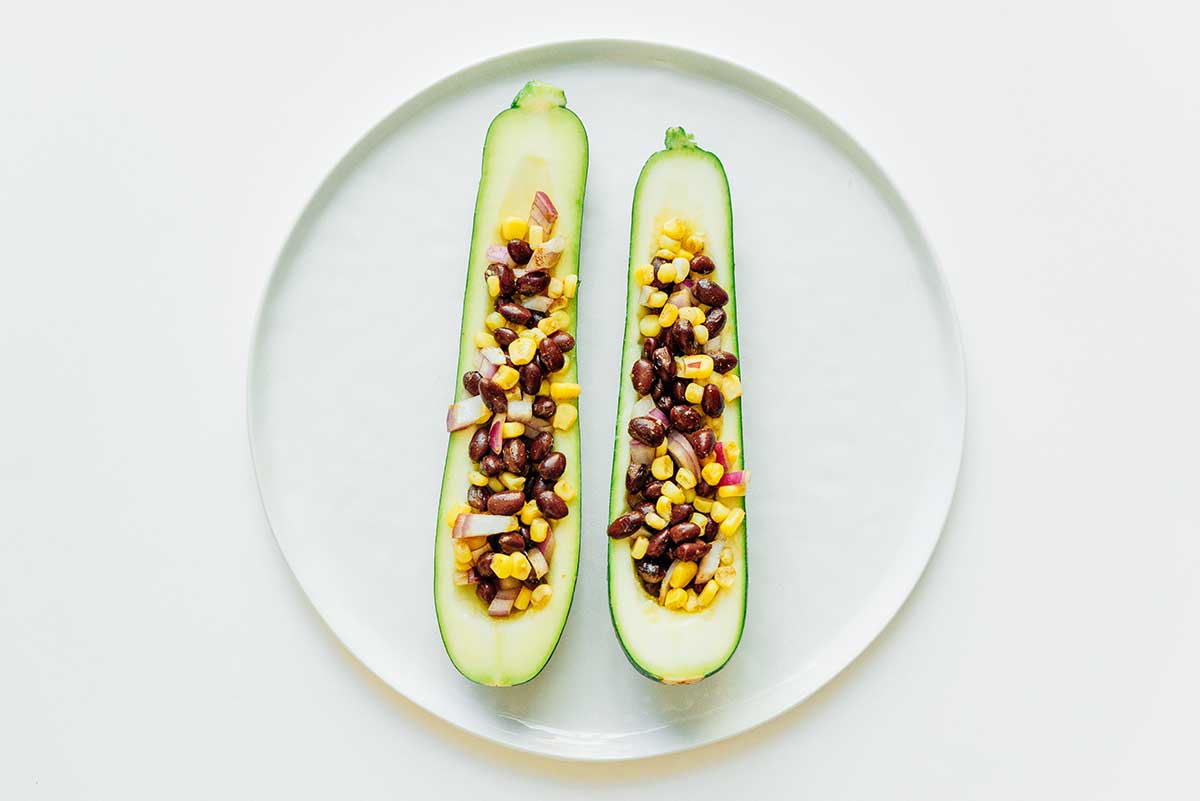 Two uncooked stuffed zucchini halves on a white plate filled with corn, black beans, cumin, and diced red onion