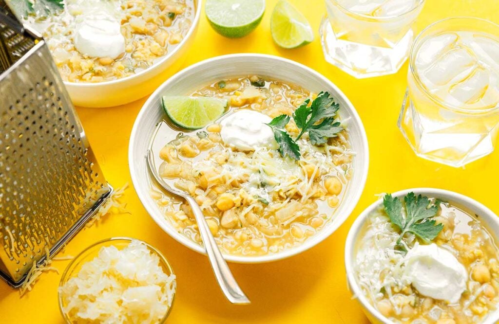 A white bowl filled with tempeh white bean chili and garnished with cilantro, Greek yogurt, shredded cheese, and a slice of lime
